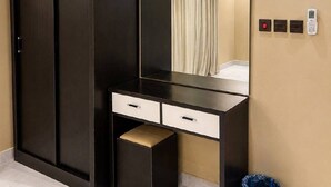 Een kamer met een garderobe, een bureau met lades, een spiegel, een airconditioningapparaat, een prullenbak en een aan de muur bevestigd telefoon.