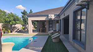 A backyard with a swimming pool, a covered patio, and a large window. - Petalite Villa Maun (Maun)
