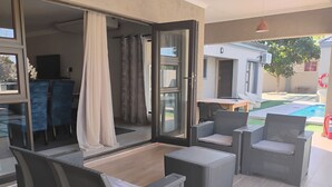 A modern outdoor living area with wicker furniture, a glass door leading to a pool, and a view of the backyard. - Petalite Villa Maun (Maun)