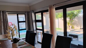A dining area with a wooden table, chairs, and a view of a swimming pool and garden through large windows. - Petalite Villa Maun (Maun)