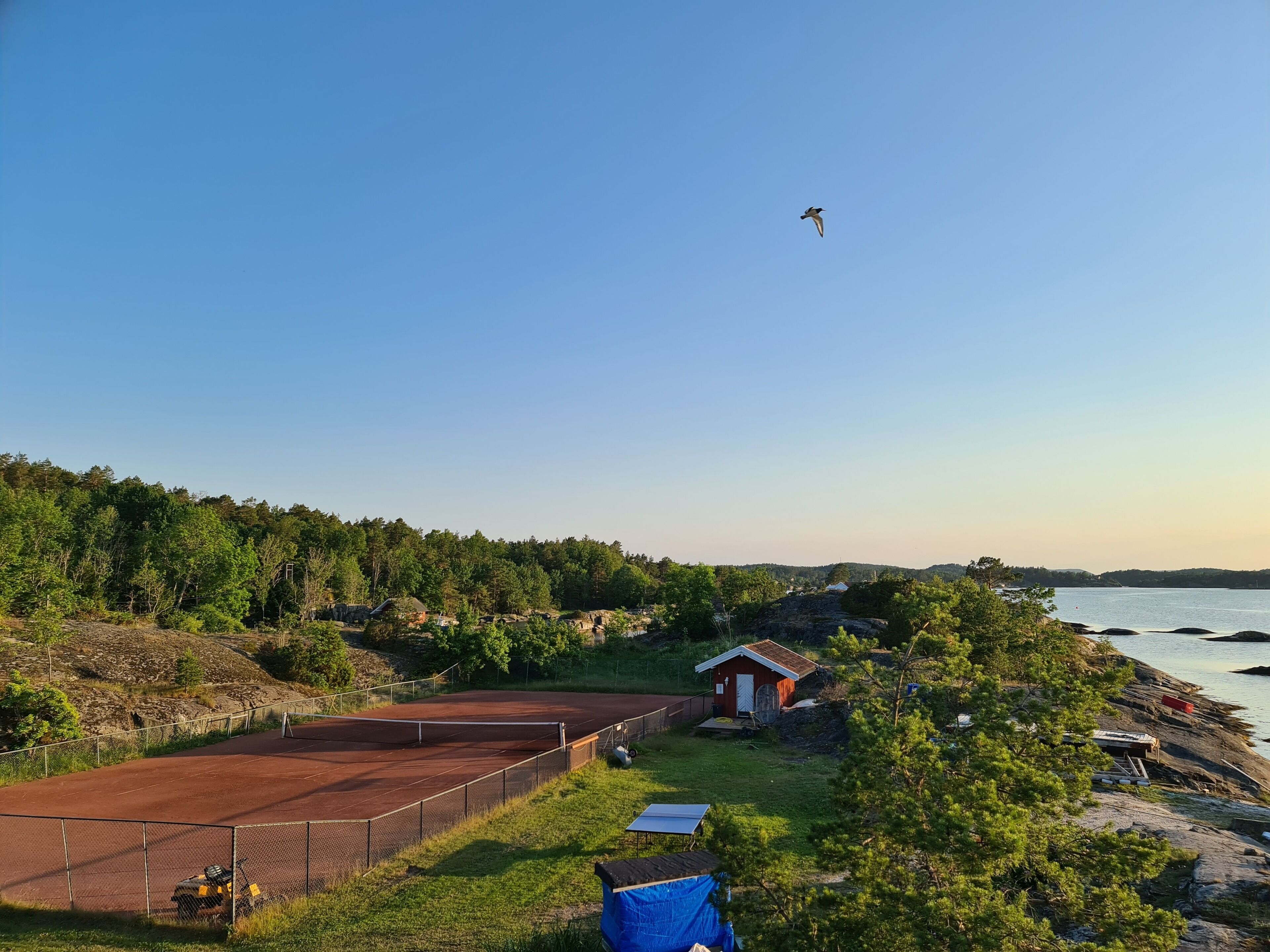 Un court de tennis avec un filet, entouré d’une clôture, situé près d’un petit bâtiment rouge et d’une étendue d’eau.