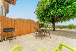 Un patio con cerco de madera, una mesa con sillas, una parrilla y un árbol.