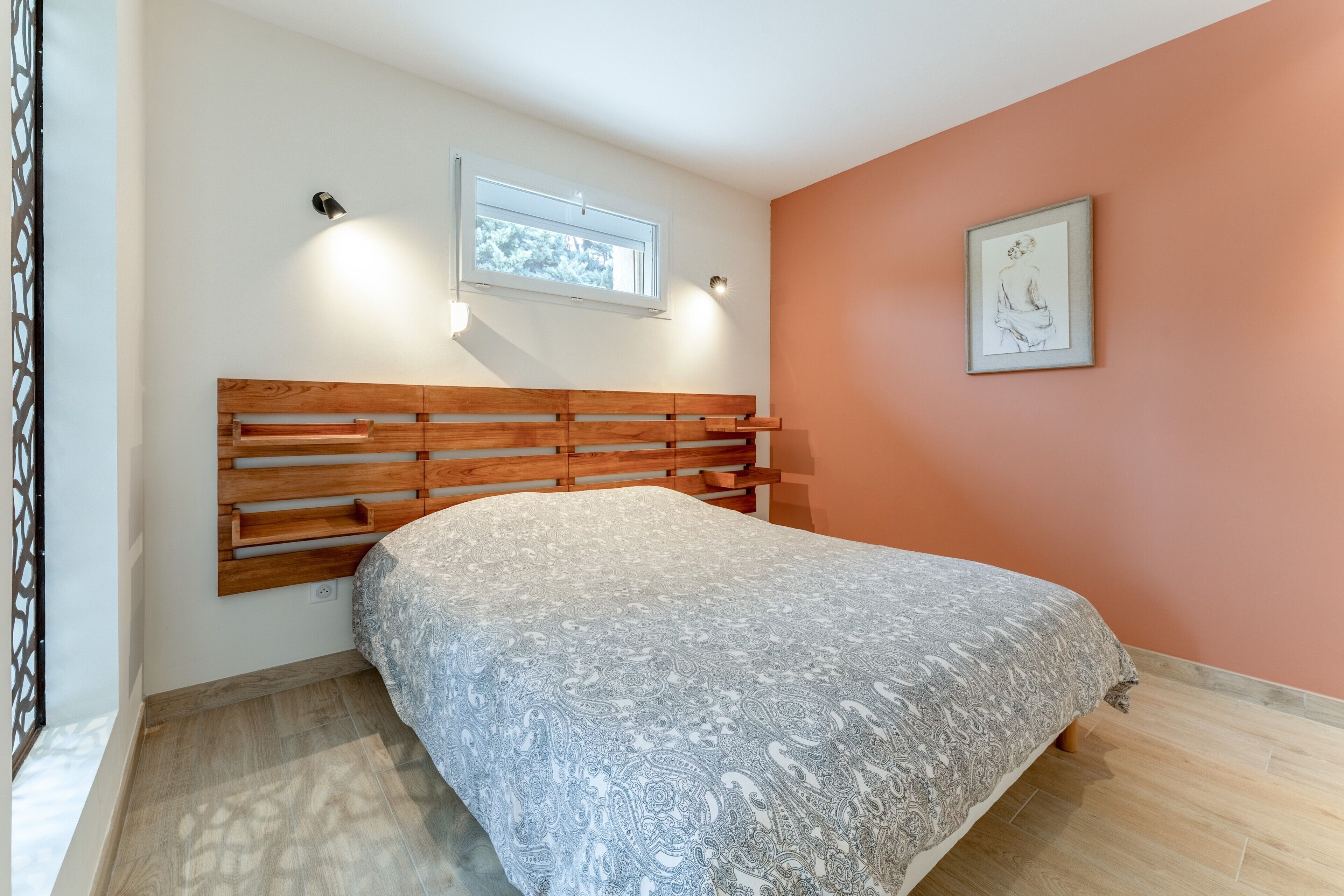 A bedroom with a wooden headboard, a patterned bedspread, a window, and a framed picture on the wall.