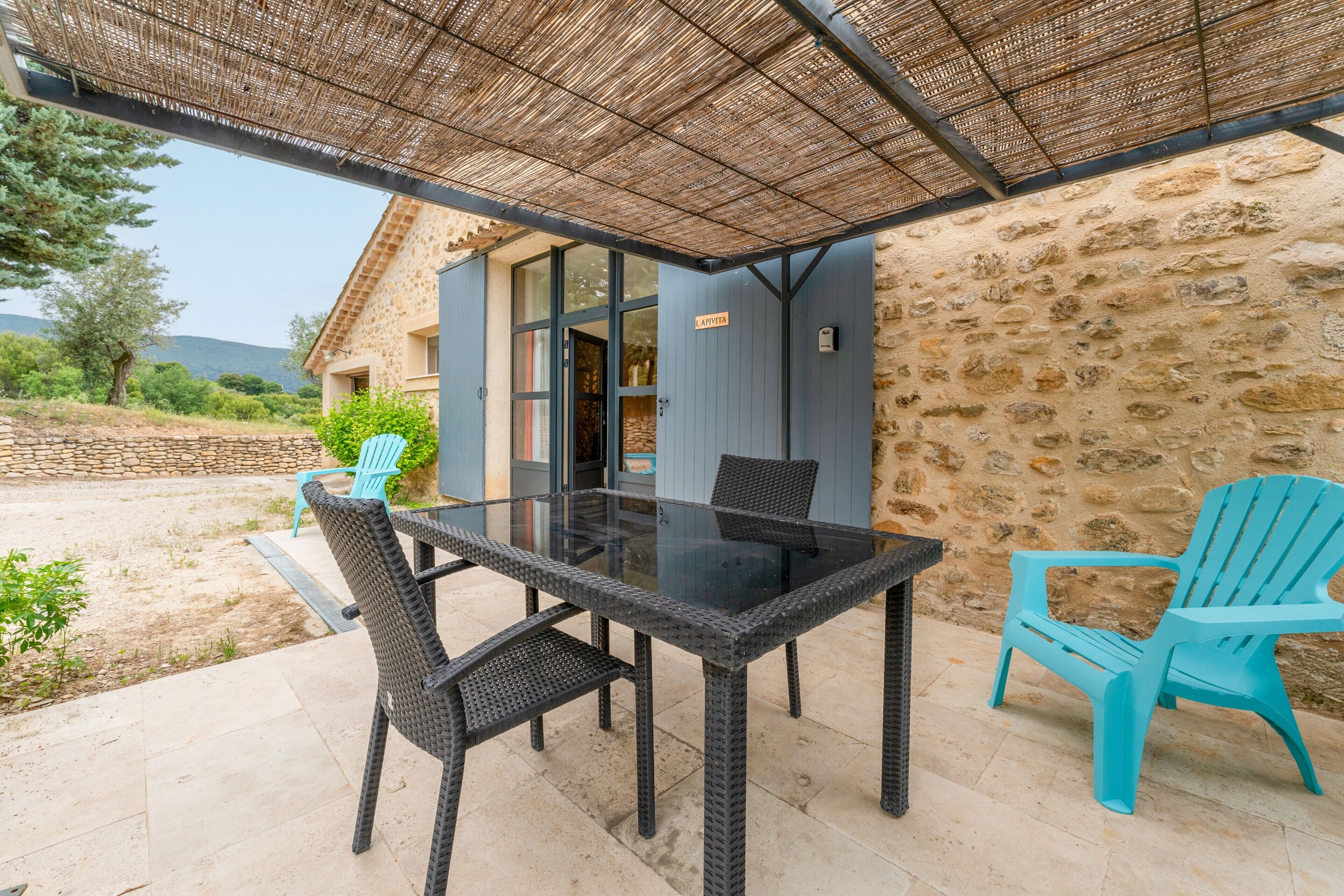 A covered outdoor dining area with a stone wall, a glass table, wicker chairs, and a blue chair.