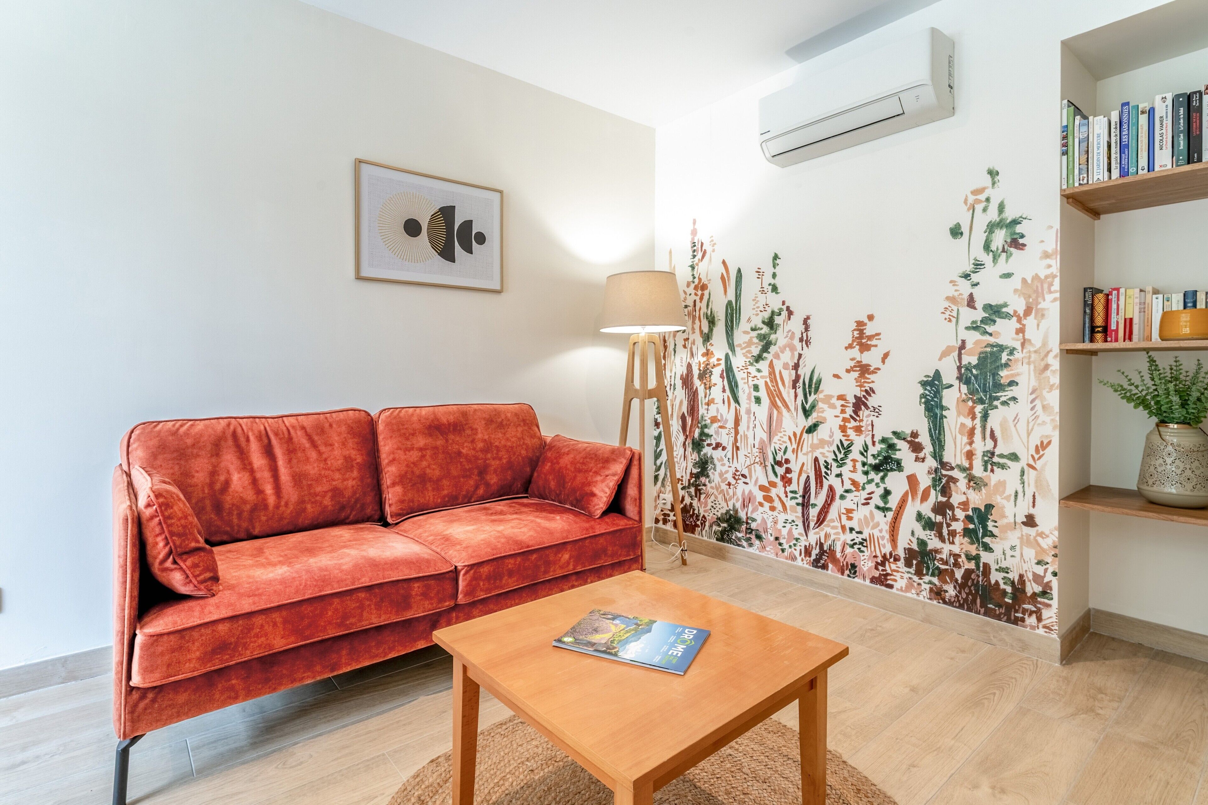 A living room with a red sofa, a wooden coffee table, a floor lamp, a bookshelf, and a wall with a floral mural.