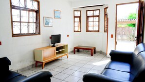 A living room with a TV on a wooden cabinet, a bench, and two black sofas. - Casa com ótima localização e churrasqueira (Ilhabela)