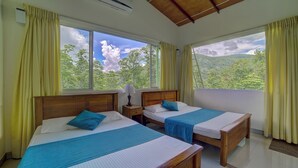 Ein Zimmer mit zwei Betten, einem Nachttisch, einer Lampe und Blick auf eine bewaldete Landschaft durch große Fenster.