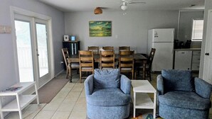 Ein Wohnzimmer mit zwei blauen Sesseln, einem Esstisch mit Holzstühlen, einem weißen Kühlschrank und einem Deckenventilator.