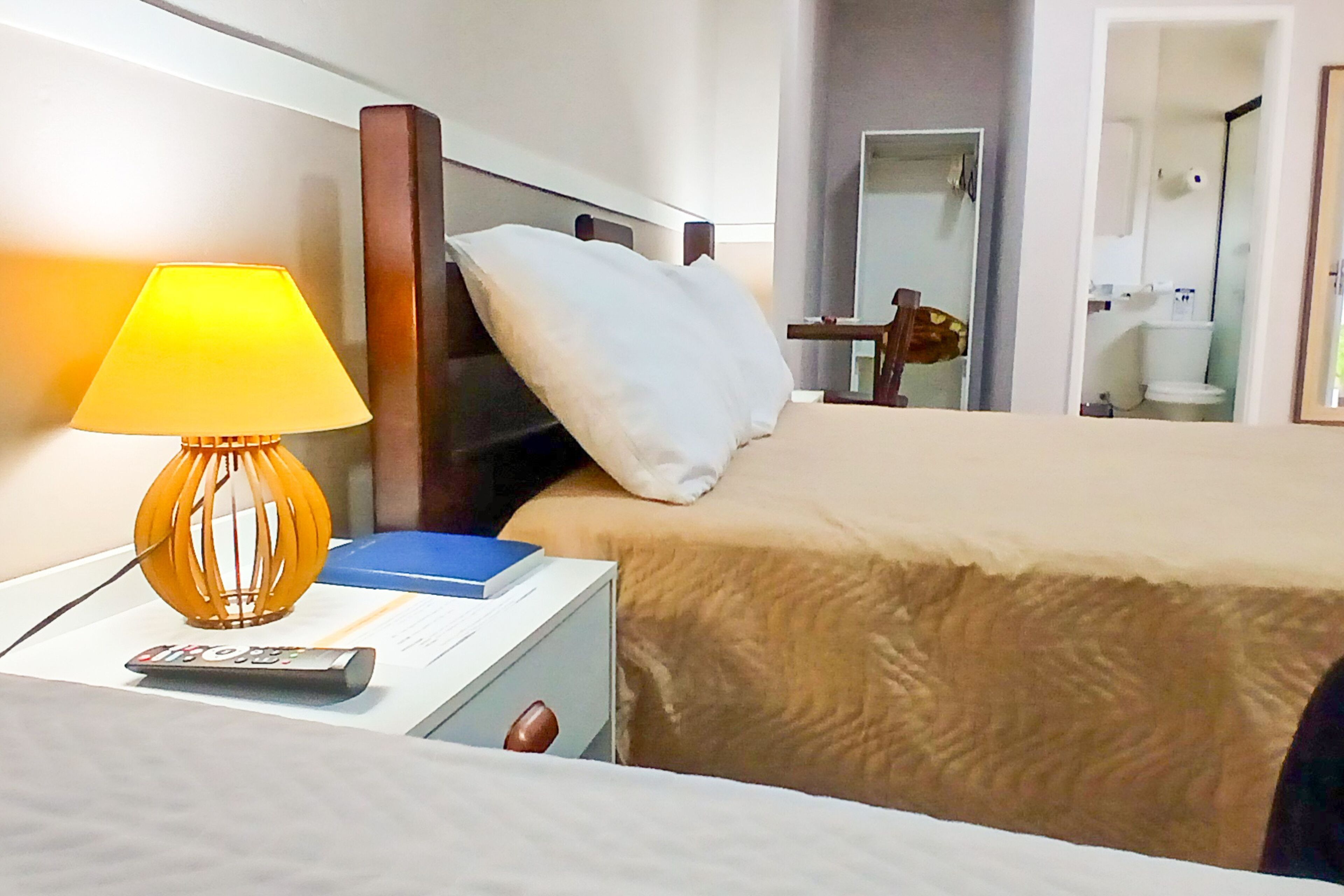 A hotel room with a bed, a nightstand with a lamp, a remote control, and a blue book.