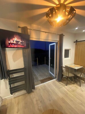A room with a TV displaying a fireplace, a balcony with a wooden floor, and a dining area with chairs and a table. - Hill View Cabin-private hot Tub-valley Views (Knighton)