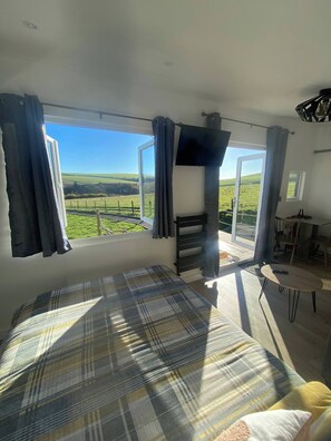A bedroom with a large bed, a small table, and a view of a green landscape through a glass door. - Hill View Cabin-private hot Tub-valley Views (Knighton)