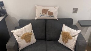 A grey sofa with two white pillows featuring a brown dog design and a central pillow with a similar design and text. A round wooden coffee table is in front of the sofa. - Hill View Cabin-private hot Tub-valley Views (Knighton)