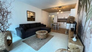 A modern living room with a dark sofa, a round coffee table, and a wooden side table with a vase. A kitchen is visible in the background with white cabinets and a dining area. - Apartment in Grau du Roi by the Beach (Le Grau-Du-Roi)