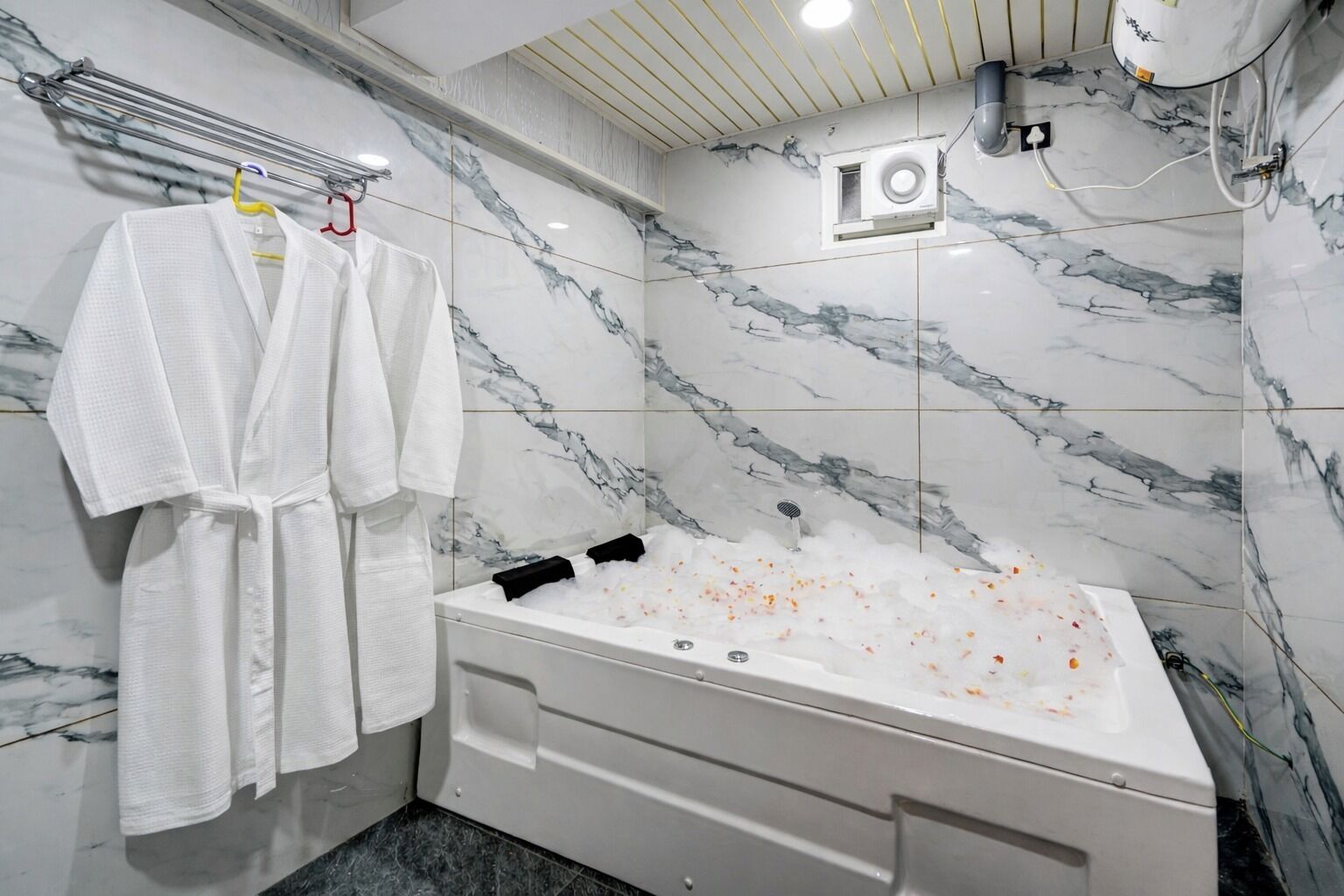 Une salle de bain avec un mur en marbre, une baignoire remplie de mousse, et deux peignoirs blancs suspendus à un porte-serviettes.
