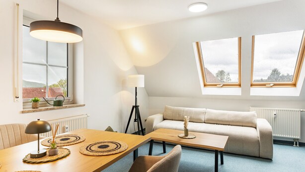 Un salon moderne avec un canapé, une table à manger avec des sets de table, une lampe et un lampadaire. Il y a des fenêtres de toit et une vue sur l’extérieur.