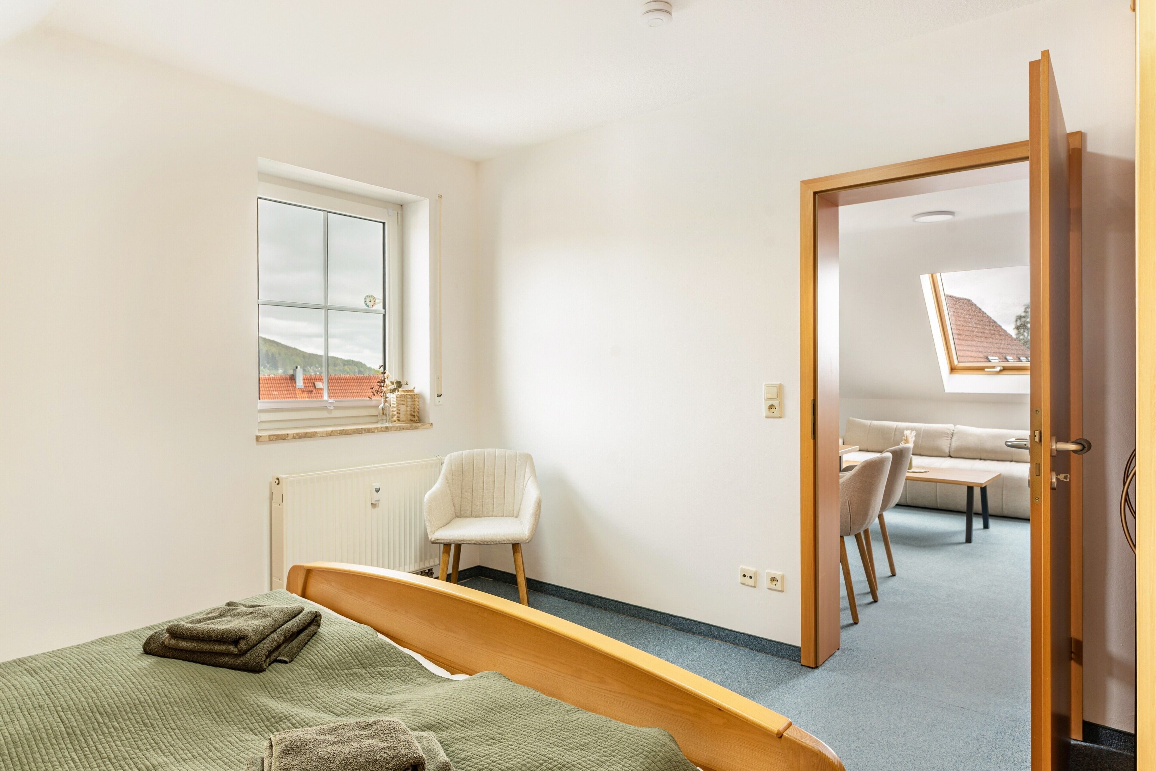 A bedroom with a bed, a chair, a window, and a view of a dining area.