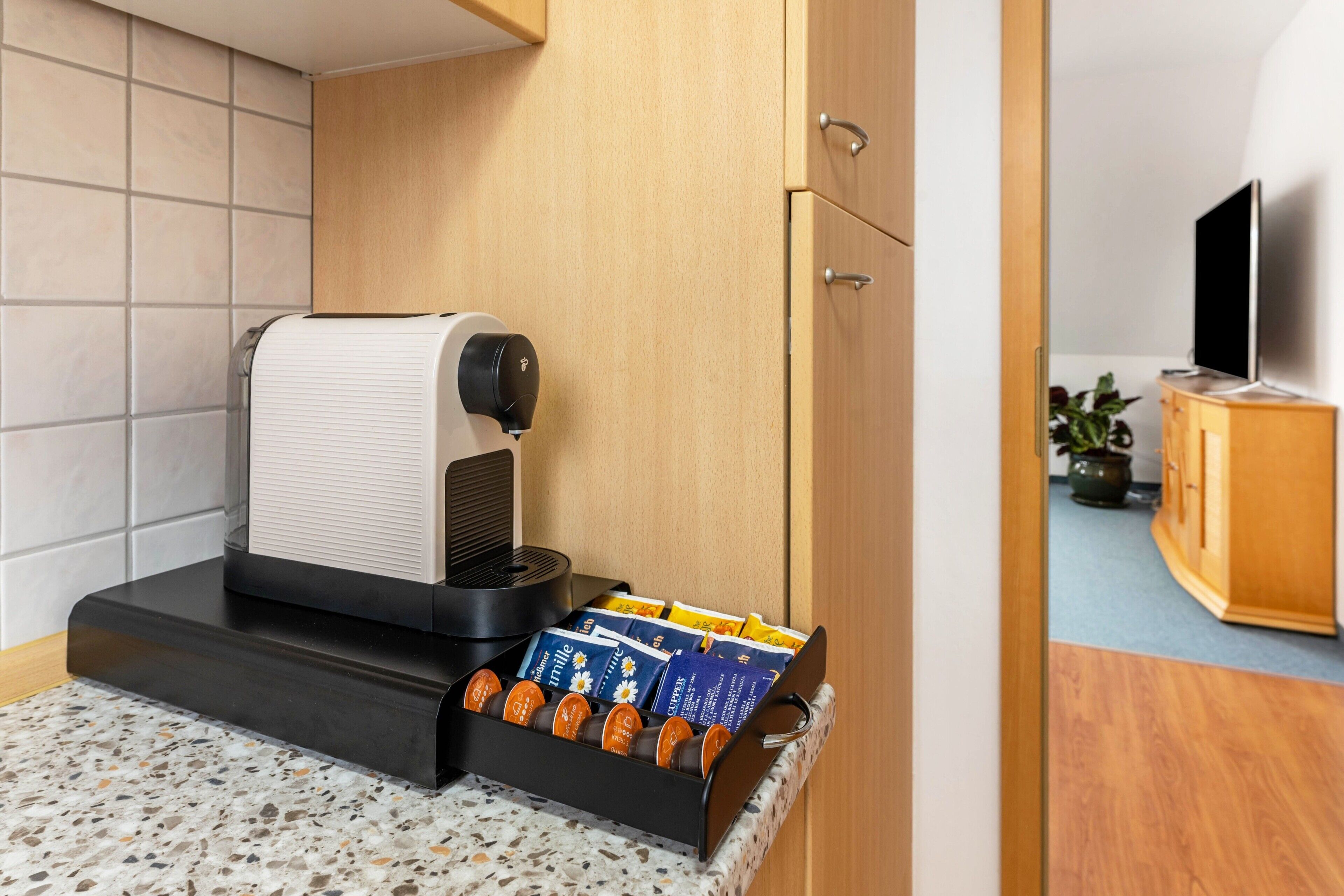 Eine Kaffeemaschine mit Kapselhalter, ein Fernseher und eine Holzkommode in einem Zimmer mit gefliesten Wänden.