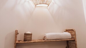 Casa de banho com prateleira de fibra natural com rolo de papel higiénico e toalhas de papel, uma cadeira de vime e um candeeiro fixo na parede.
