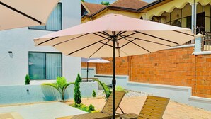 A poolside area with lounge chairs and a large umbrella, surrounded by a modern building with a tiled patio and a brick wall. - 2-bedroom apartment in Kigali with AC, a swimming pool, and great city views. (Kigali)