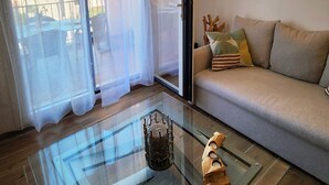 Un salón moderno con una mesa de centro de vidrio, un sofá con cojines y vistas al exterior a través de cortinas de gasa.