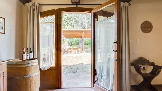 Une porte ouverte donne sur un patio extérieur, où se trouvent une table et des chaises en bois, un sol en pierre et un tonneau en bois contenant des bouteilles.