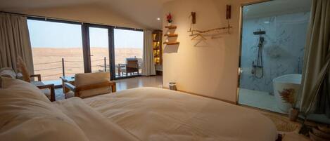Een ruime slaapkamer met een groot bed, een zithoek met een tafel, en uitzicht op de woestijn door grote ramen.