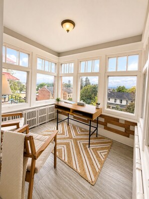 A corner room with a desk, chairs, a radiator, and large windows with a view of buildings and trees.