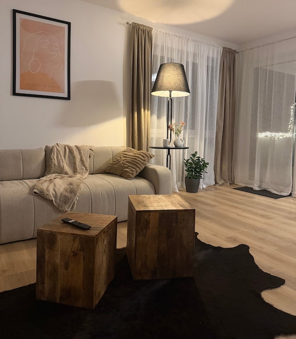 Un salon comprenant un canapé, deux petites tables en bois, une lampe, un tableau encadré et une plante en pot.