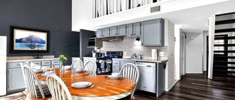A modern kitchen with a dining area, a TV mounted on the wall, and a staircase leading to an upper level.