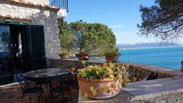 Un bâtiment en pierre doté d’une terrasse qui donne sur un plan d’eau. On y trouve une table, des chaises et des plantes en pot.