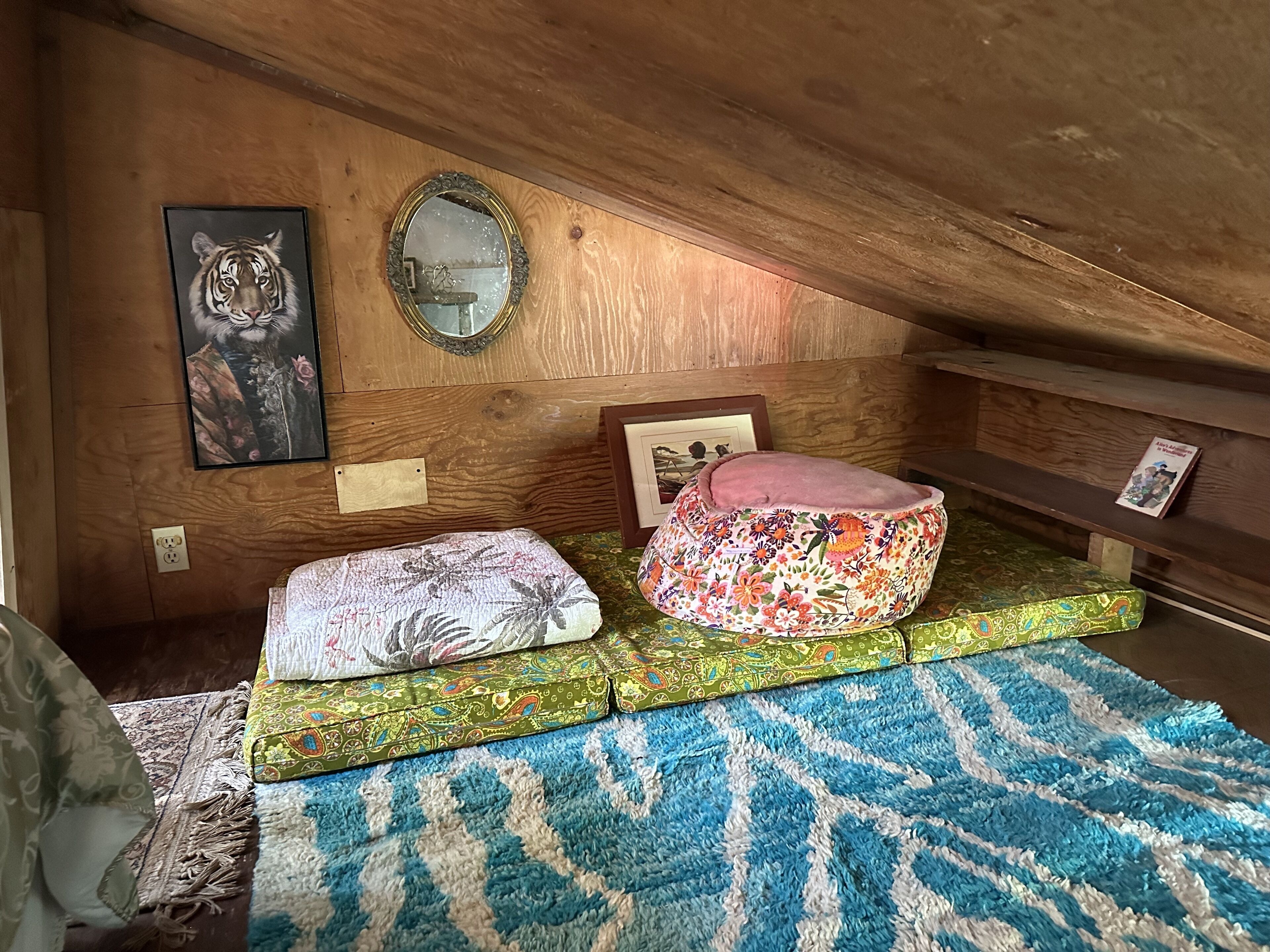 Un dormitorio acogedor en el ático, con techo inclinado de madera, alfombra azul y blanca estampada, dos camas coloridas para mascotas, un cuadro enmarcado de un tigre y un espejo.