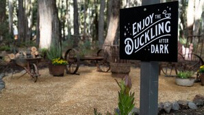 A sign with the text 'ENJOY THE DUCKLING AFTER DARK' in a garden setting with trees, a wheelbarrow, and potted plants. - The Duckling quintessential bush retreat set amongst towering gums. One bedroom - dog friendly (Wheatsheaf)