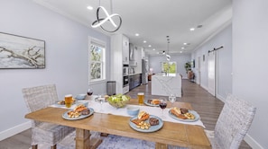 A modern dining area with a wooden table set for a meal, a kitchen in the background, and a view of the outdoors. - Beach Getaway - Ask about Special Monthly Rates (Hernando Beach)
