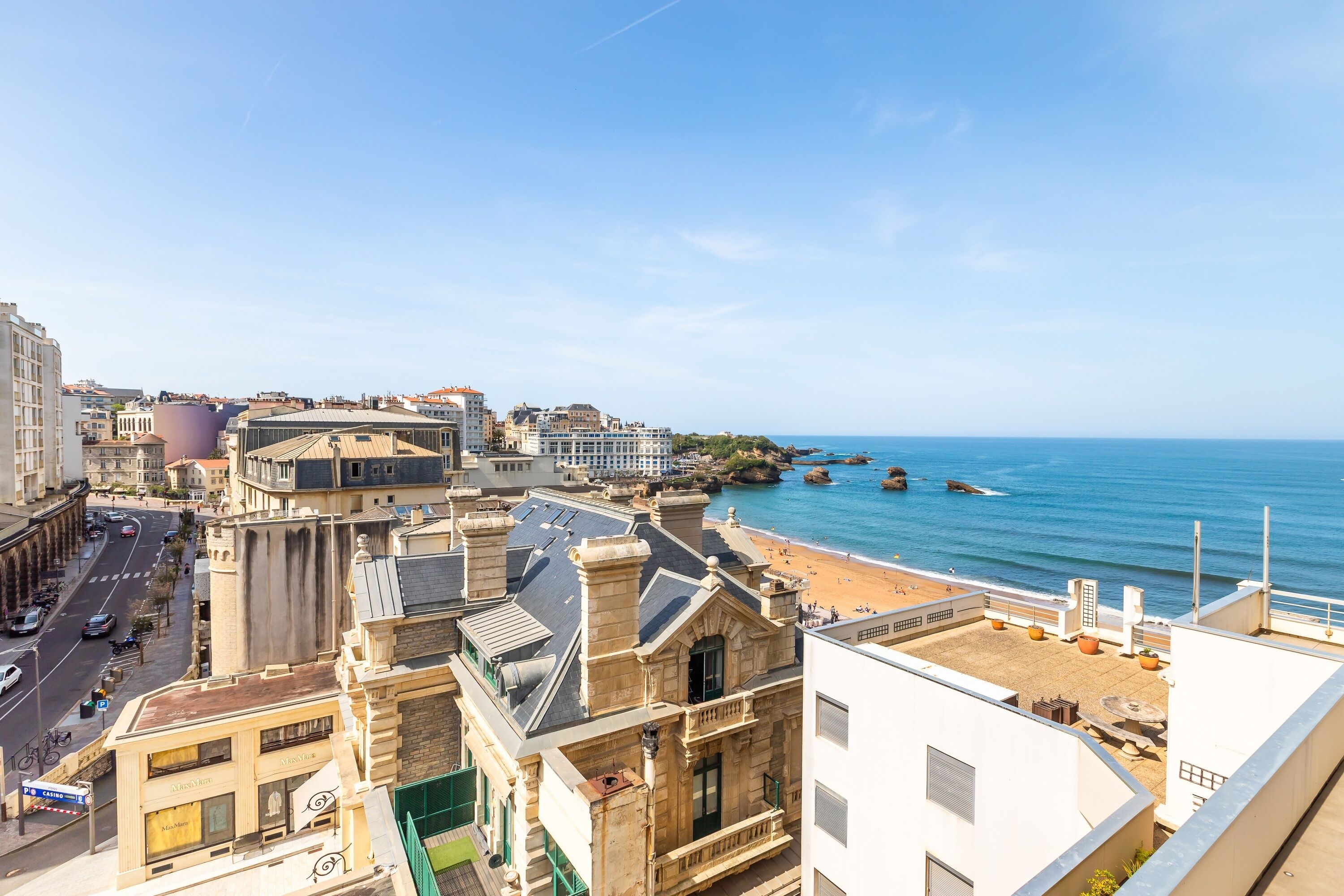A coastal cityscape with a prominent building featuring a rooftop terrace overlooking the beach.