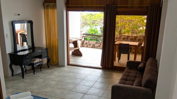 Une chambre d’hôtel comprenant un lit, un canapé, une commode, un miroir et donnant sur une terrasse extérieure avec une table et des chaises.