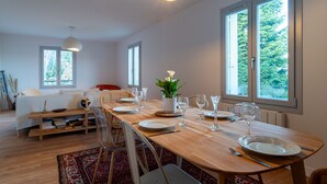 Uma área de refeições com mesa de madeira posta para quatro pessoas, cercada por cadeiras. Há janelas amplas que proporcionam luz natural e vista para o verde lá fora.