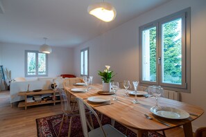 Ein Essbereich mit einem Holztisch, gedeckt für vier Personen, umgeben von Stühlen. Große Fenster sorgen für natürliches Licht und bieten Blick ins Grüne.