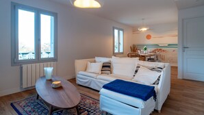 Uma sala de estar moderna com um sofá branco, uma mesa de centro de madeira e um espaço de refeições com mesa posta para dois.