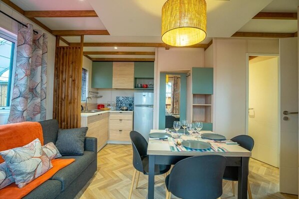 A modern kitchen with wooden beams, a dining area with a table set for four, and a cozy living area with a sofa and cushions. - Camping la Clairière (Rouffilhac)