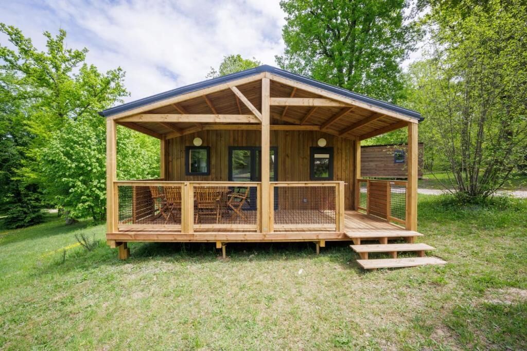 Una cabaña de madera con porche y un área cercada, rodeada de vegetación.