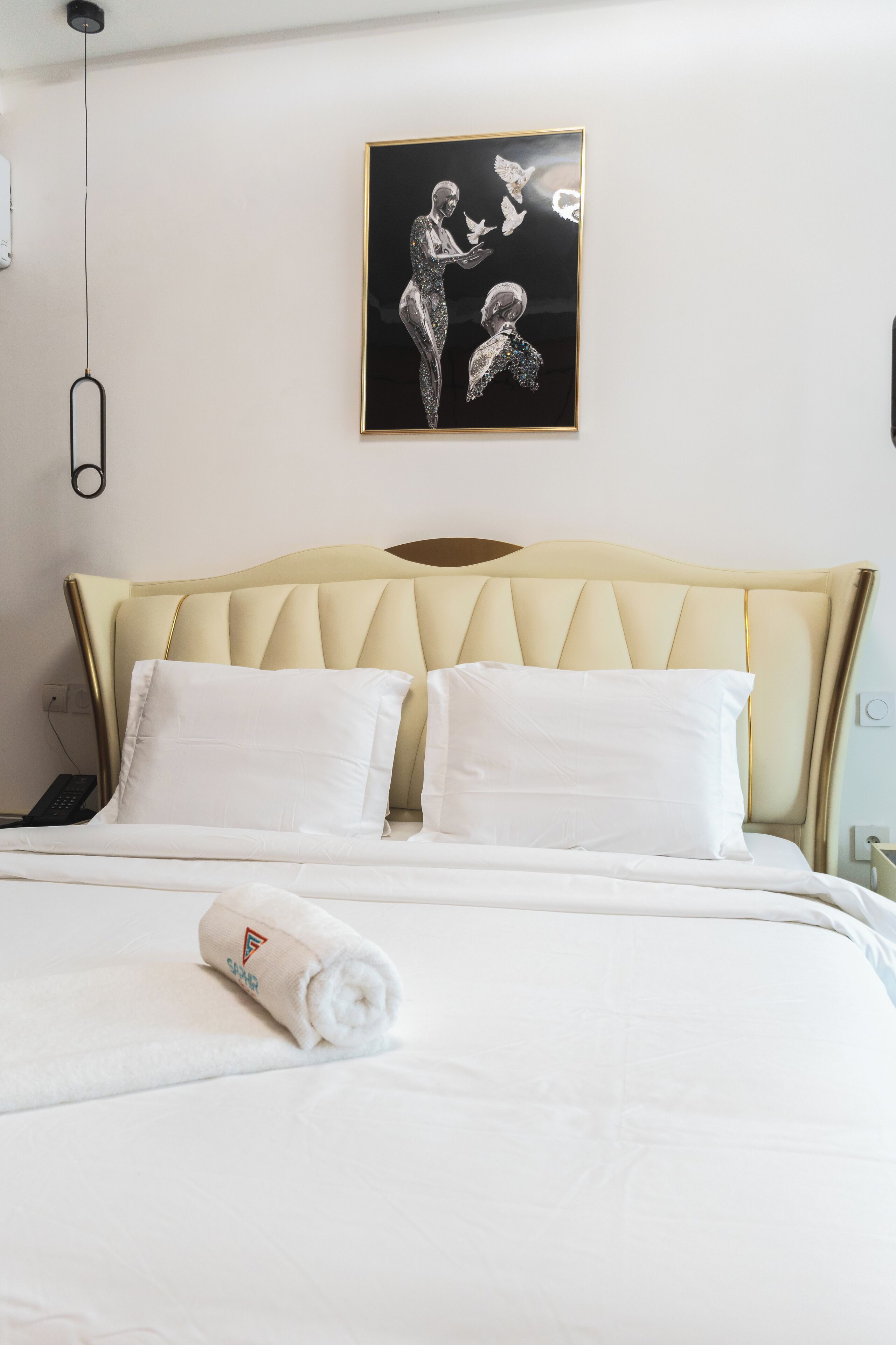 A neatly made bed with white linens and pillows, a cream-colored headboard, and a framed artwork on the wall.