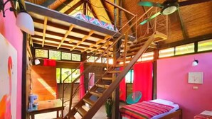 A room with a wooden staircase, a bed with a colorful blanket, and a dining area with a table and chairs. - Pizote - Jungle Cabina with Pool Access (Cabuya)