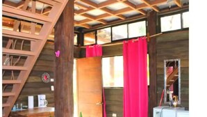 A rustic cabin interior with a wooden staircase, a dining area, a kitchen with a white refrigerator, and a large window with red curtains. - Pizote - Jungle Cabina with Pool Access (Cabuya)