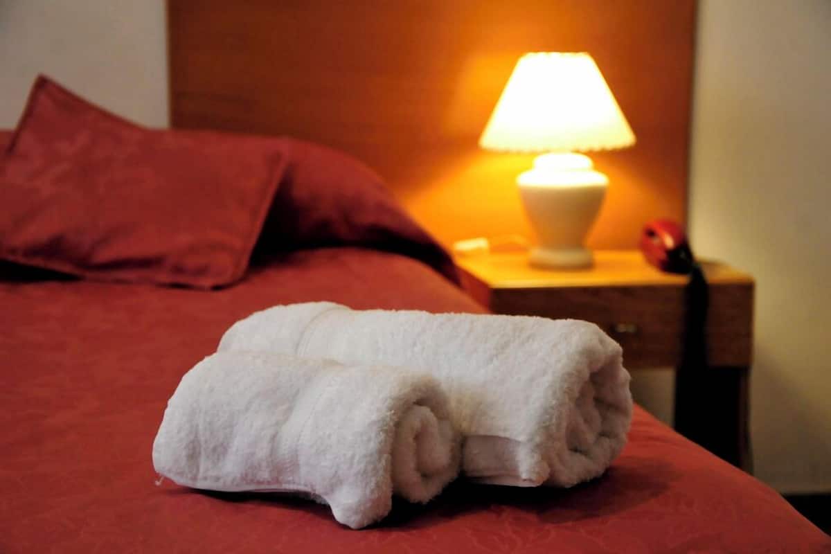 Un lit bien fait, avec une serviette blanche roulée placée au centre, un oreiller rouge et une lampe de chevet posée sur une table de nuit en bois.