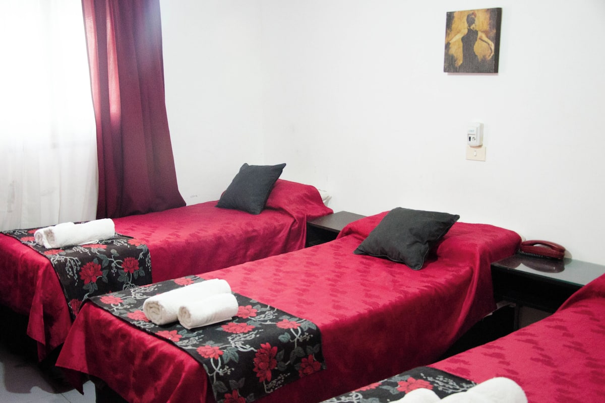 Une chambre d’hôtel avec deux lits simples, chacun recouvert d’une couverture rouge et agrémenté d’un coussin à motifs floraux noirs. Une petite table se trouve entre les lits, avec un téléphone et une prise électrique.
