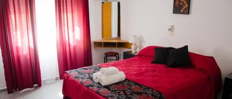 A hotel room with a red bedspread, a desk with a chair, a wall-mounted air conditioner, and a painting on the wall.