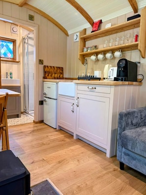 A compact kitchen with white cabinets, a coffee machine, and a wooden shelf with cups.