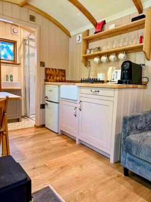 A compact kitchen with a refrigerator, a coffee maker, and a shelf with cups.