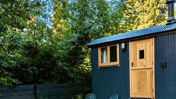 A small wooden cabin with a metal roof, a wooden door, and a window. There is a set of stairs leading to the door, and two metal chairs outside.