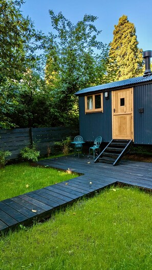 Une petite cabane en bois avec un toit en métal, une porte en bois et une fenêtre. Un escalier mène à la porte, et deux chaises en métal sont placées à l’extérieur.